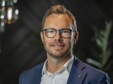 Ein Mann mit blauem Anzug und weißem Hemd. Er trägt eine Brille mit schwarzem Rahmen und einen kurzen Bart.