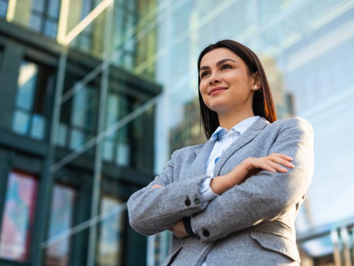 Eine junge Frau in Hemd und Blazer steht mit verschränkten Armen und selbstbewusstem Lächeln vor einem gläsernen Bürogebäude.