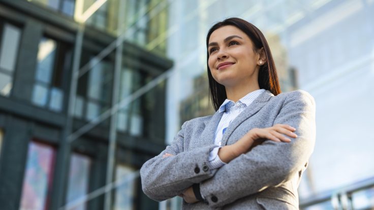 Eine junge Frau in Hemd und Blazer steht mit verschränkten Armen und selbstbewusstem Lächeln vor einem gläsernen Bürogebäude.