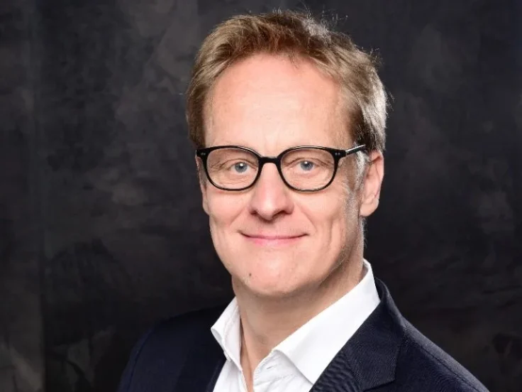 Ein Mann im Anzug lächelt. Er trägt eine schwarze Brille.