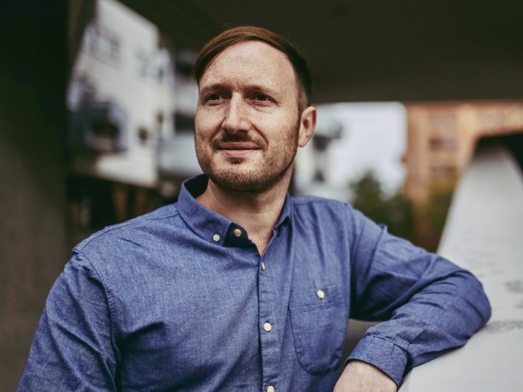 Ein Mann mit roten Haaren und blauem Hemd lehnt sich lässig an ein Geländer.