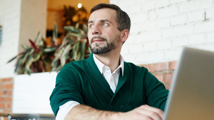 Ein Mann mit kurzen braunen Haaren und Bart sitzt an seinem Laptop und schaut nachdenklich zur Seite.