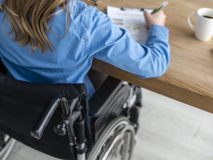 Eine blonde Frau mit hellblauem Hemd sitzt an einem Rollstuhl am Tisch. Sie füllt ein Formular aus. Neben ihr steht eine Kaffeetasse.
