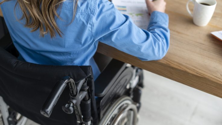 Eine blonde Frau mit hellblauem Hemd sitzt an einem Rollstuhl am Tisch. Sie füllt ein Formular aus. Neben ihr steht eine Kaffeetasse.
