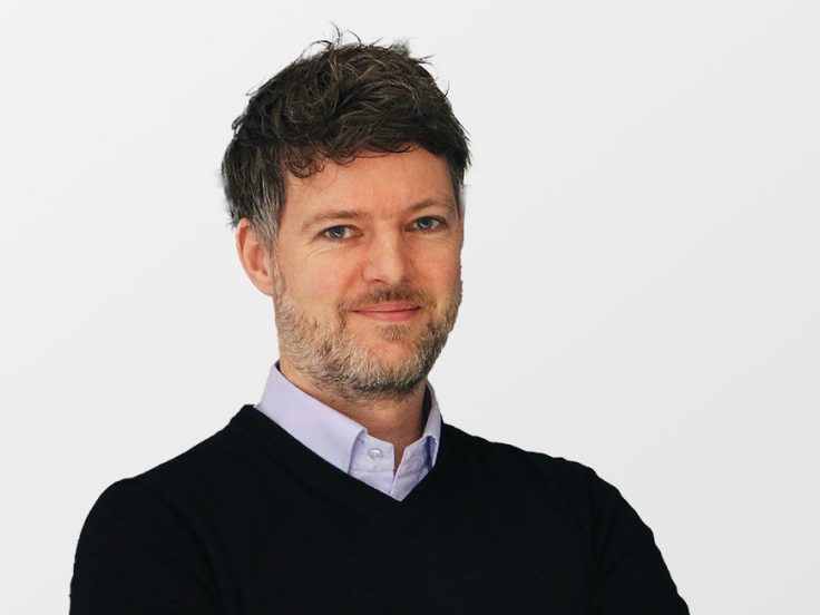 Ein Mann mit kurzem grauen Bart in Hemd und Pullover schaut in die Kamera.