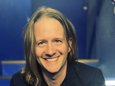 Ein Mann mit langen blonden Haaren lächelt in die Kamera.