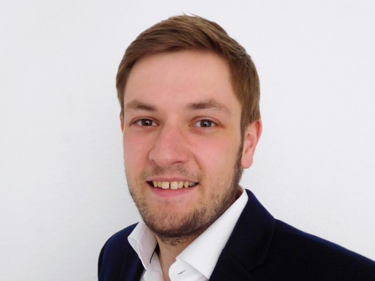 Tim Habermann steht lächelnd schräg zur Kamera gedreht. Er trägt einen Seitenscheitel, ein weißes Hemd und einen dunkelblauen Anzug.