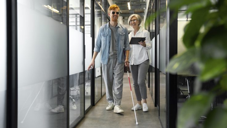 Eine sehende Frau führt einen blinden jungen Mann den Flur eines Büros entlang.