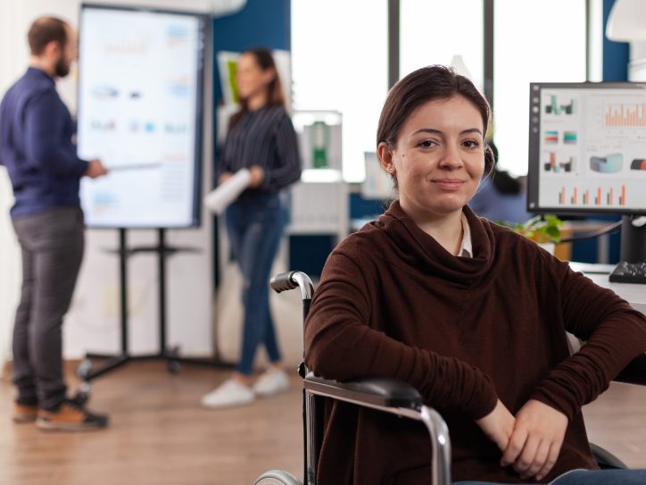Eine junge Frau in einem Rollstuhl sitzt vor ihrem Schreibtisch im Büro und lächelt. Auf ihrem Monitor sind viele Grafiken zu sehen. Im Hintergrund stehen eine Kollegin und ein Kollege vor einem Flipchart und diskutieren darüber.