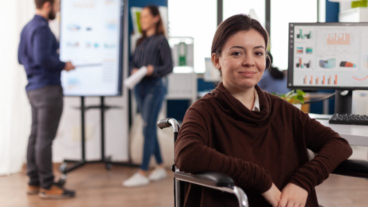 Eine junge Frau in einem Rollstuhl sitzt vor ihrem Schreibtisch im Büro und lächelt. Auf ihrem Monitor sind viele Grafiken zu sehen. Im Hintergrund stehen eine Kollegin und ein Kollege vor einem Flipchart und diskutieren darüber.