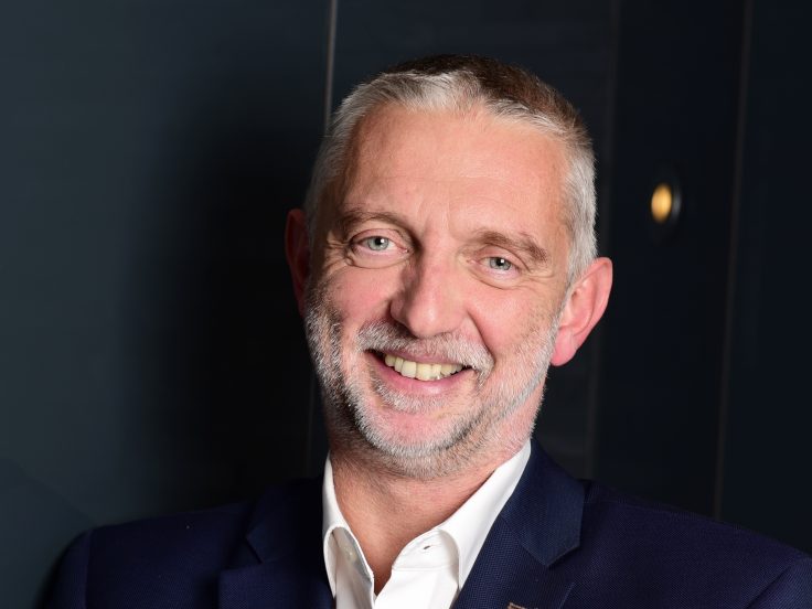 Wolfgang Schneider lächelt in die Kamera. Er graue Augen, kurze graue Haare und einen Fünf-Tage-Bart. Er trägt einen dunkelblauen Anzug und ein weißes Hemd.